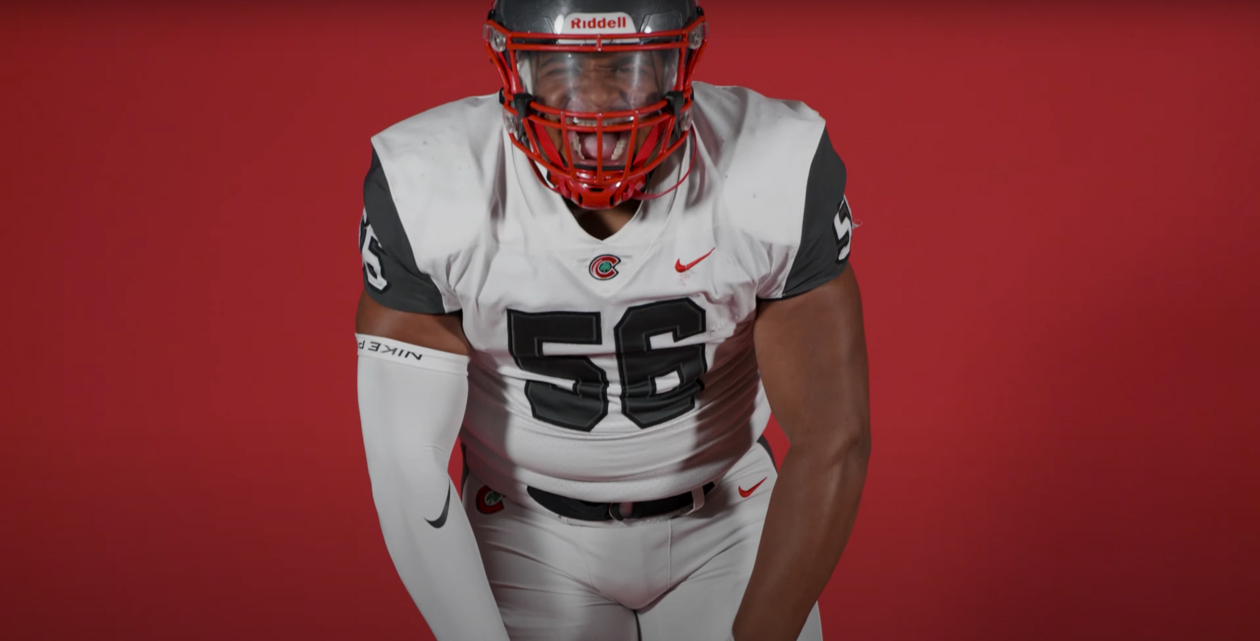 Football player wearing white jersey with number 56 against red backdrop.