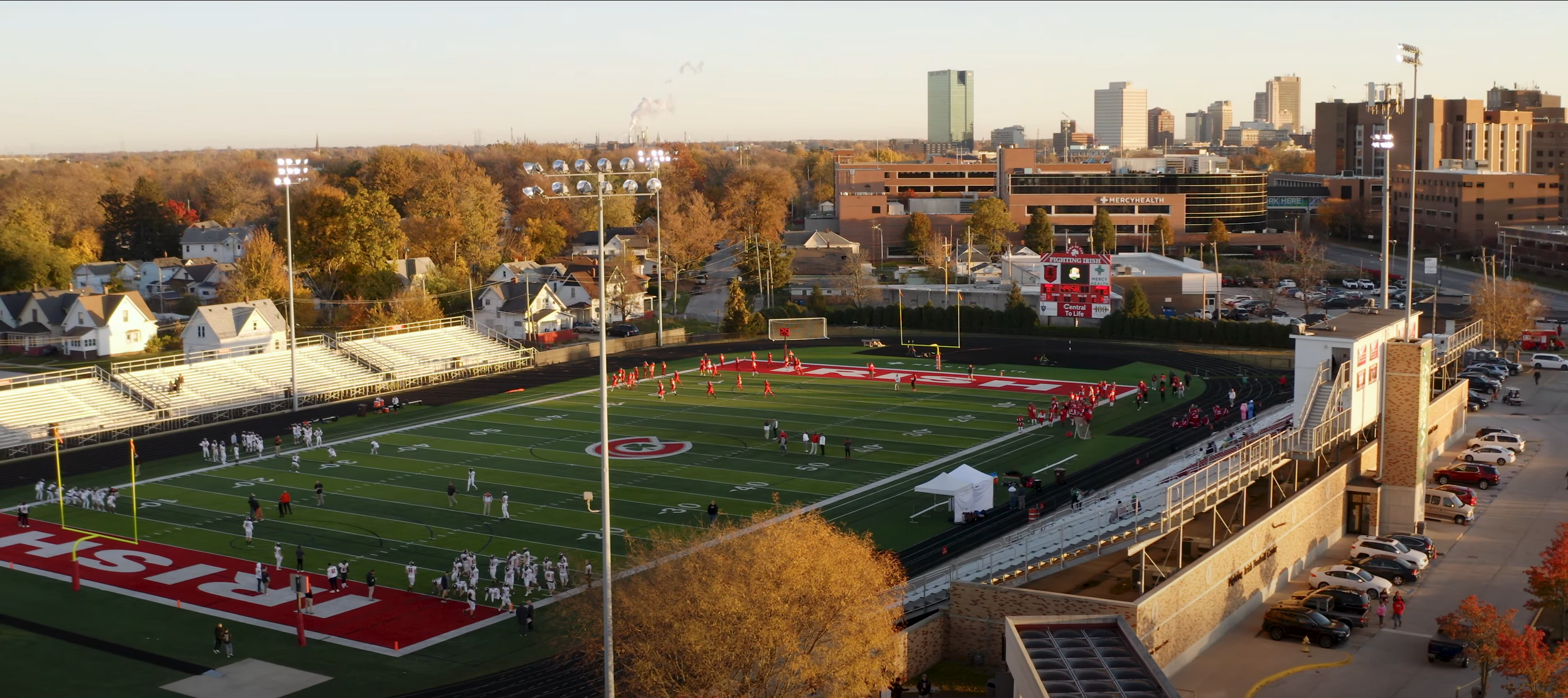 Aerial view of a sports field with artificial turf surrounded by tall buildings and autumn trees at sunset.
