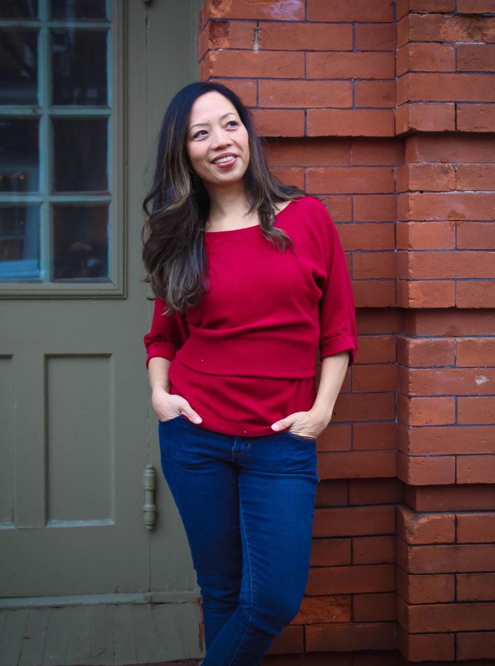 Woman in red sweater poses casually against brick wall outdoors.