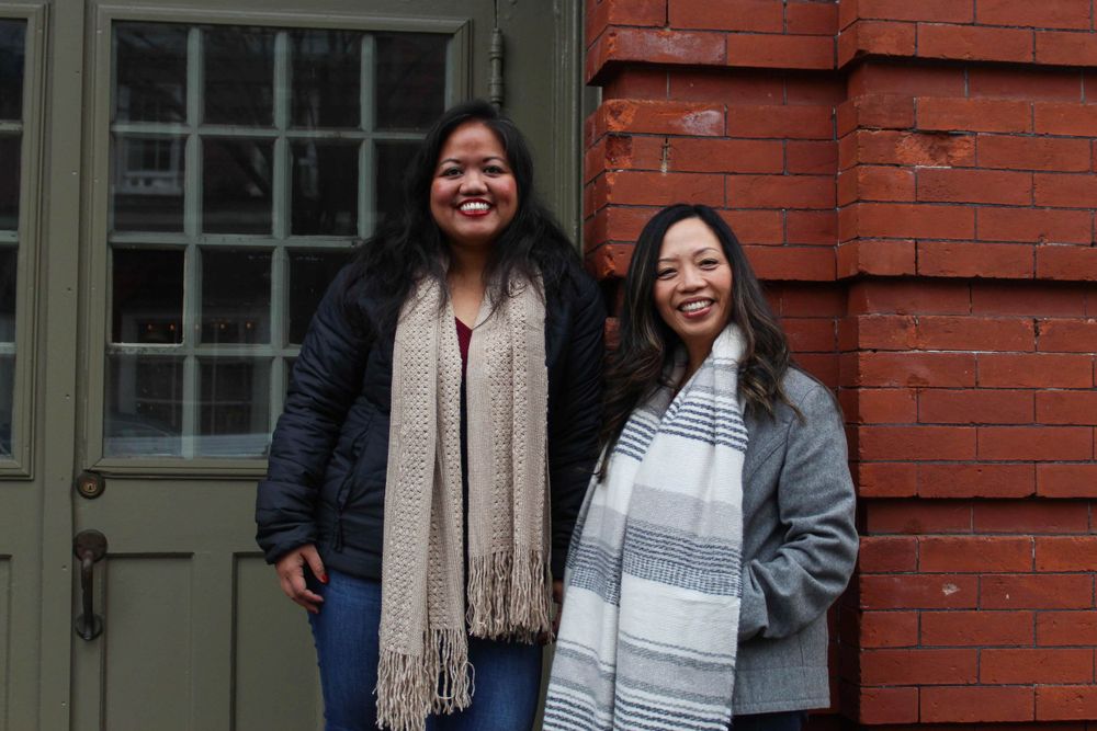 Two people standing together in winter coats and scarves outside a brick building.