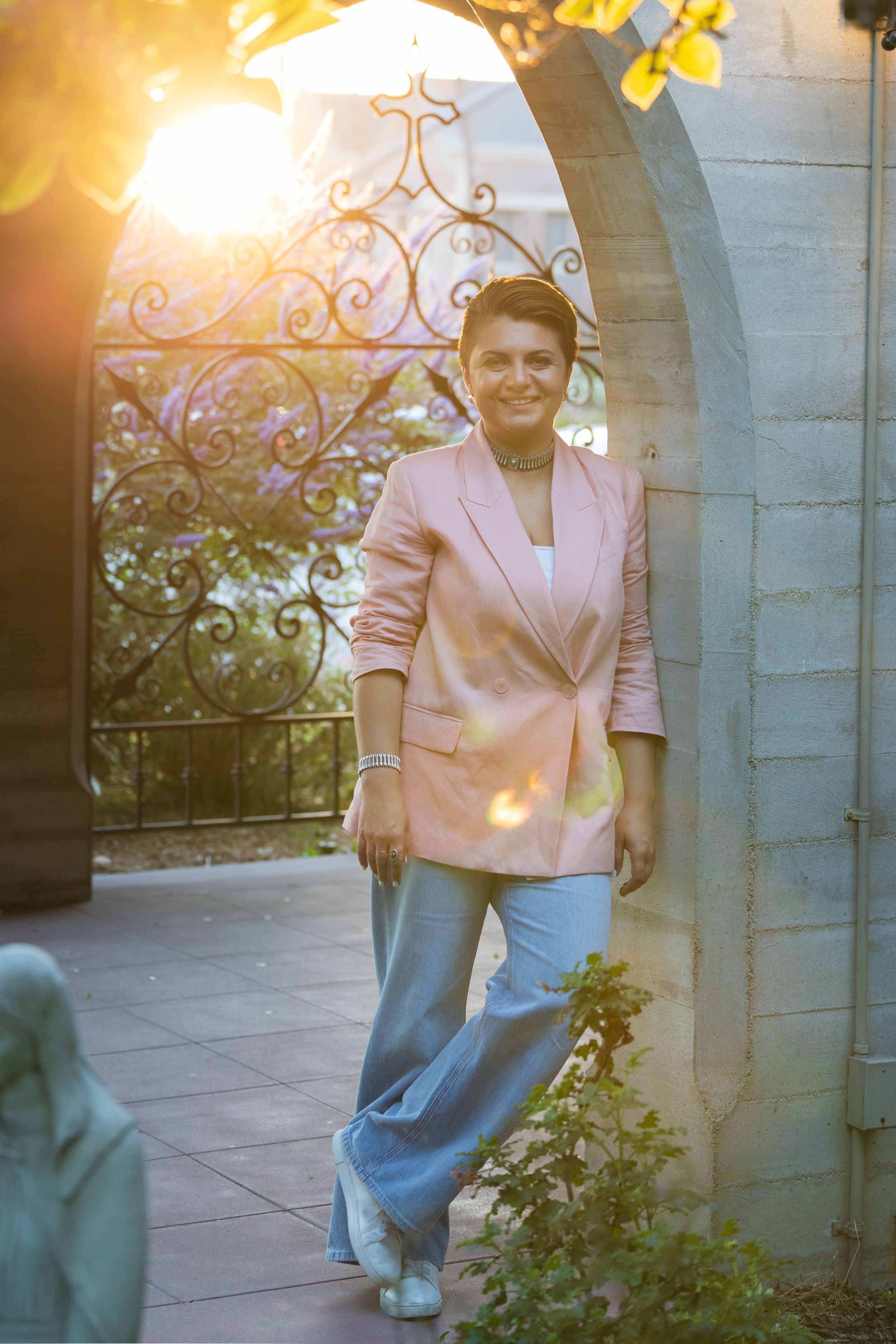 Someone poses in the evening sunlight wearing a pink blazer and jeans under an ornate stone archway with decorative ironwork.