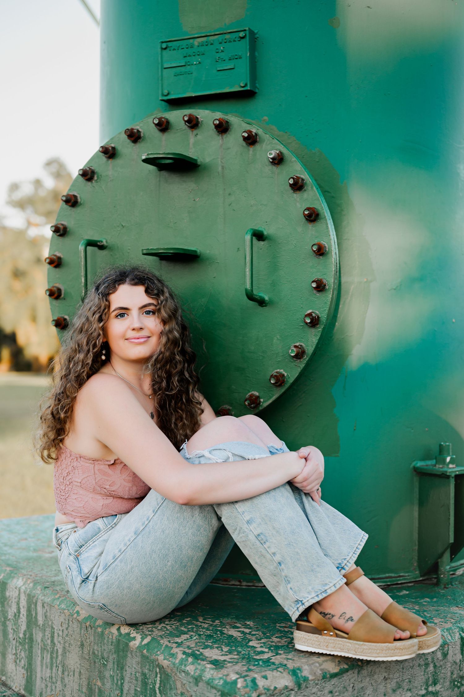 A casual portrait against a green industrial tank wearing jeans and a pink top.