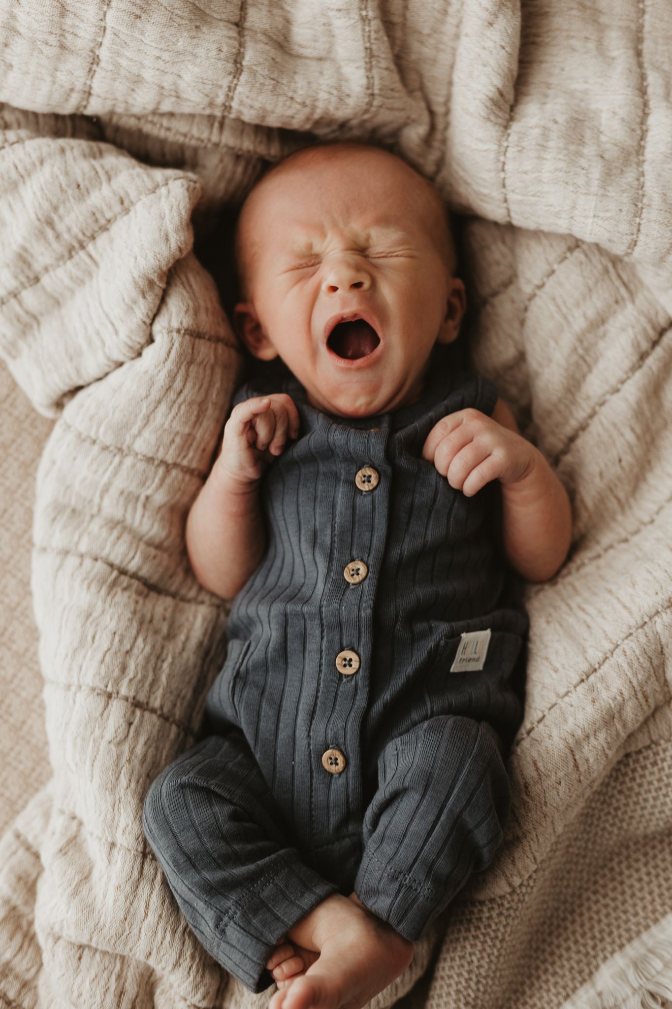 Newborn in gray buttoned romper yawning while lying on textured blanket during newborn photo session.