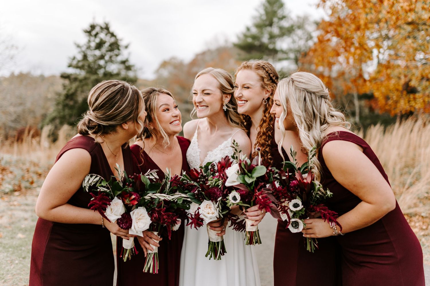 Bridesmaids in burgundy dresses share laughs with bride holding deep red and white bouquets.