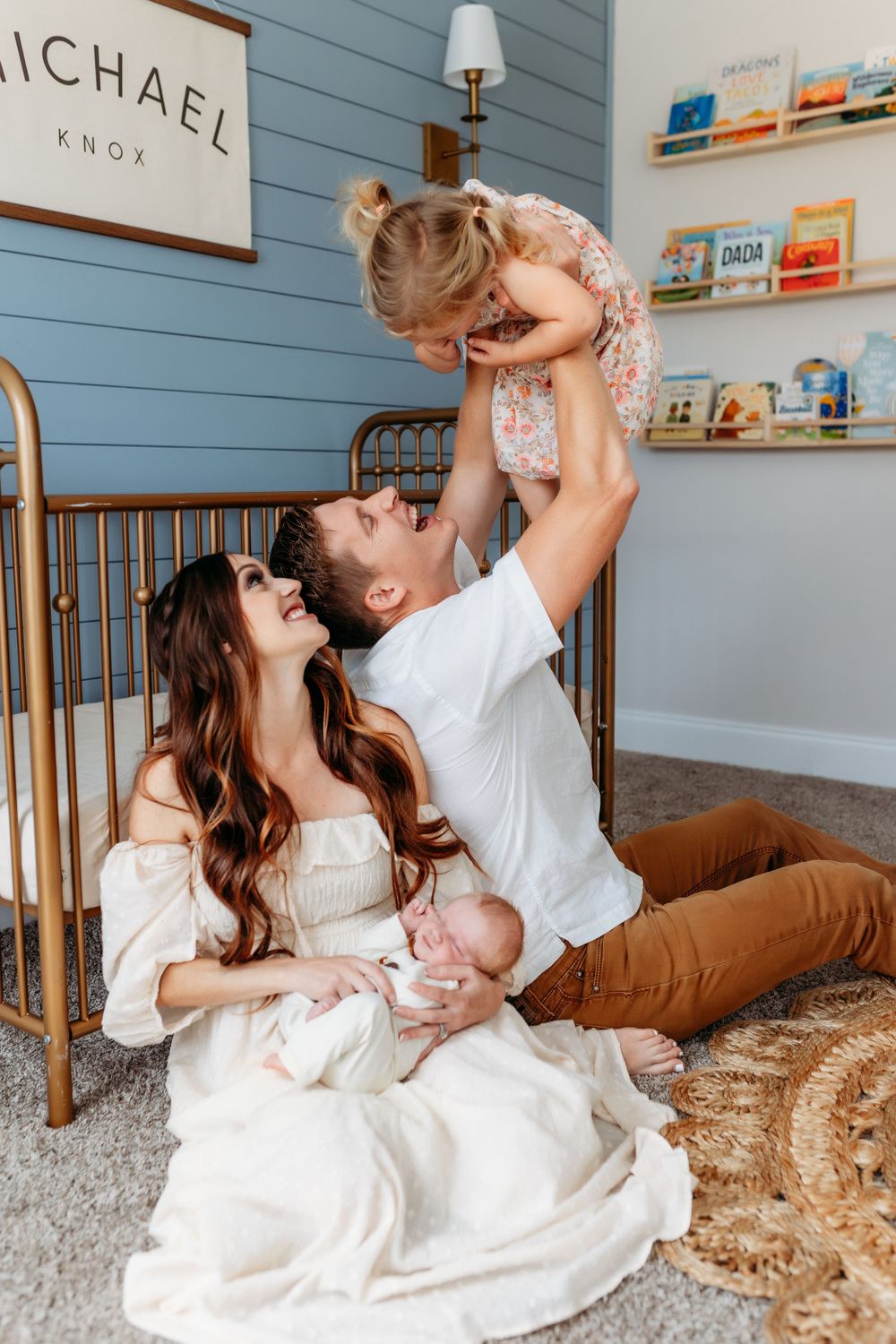 Parents sitting on nursery floor lifting toddler girl while mother holds newborn baby.
