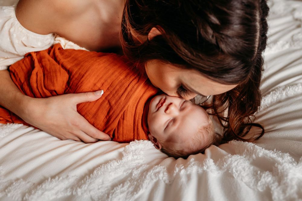 Mother tenderly kissing her newborn baby wrapped in an orange swaddle on white bedding.
