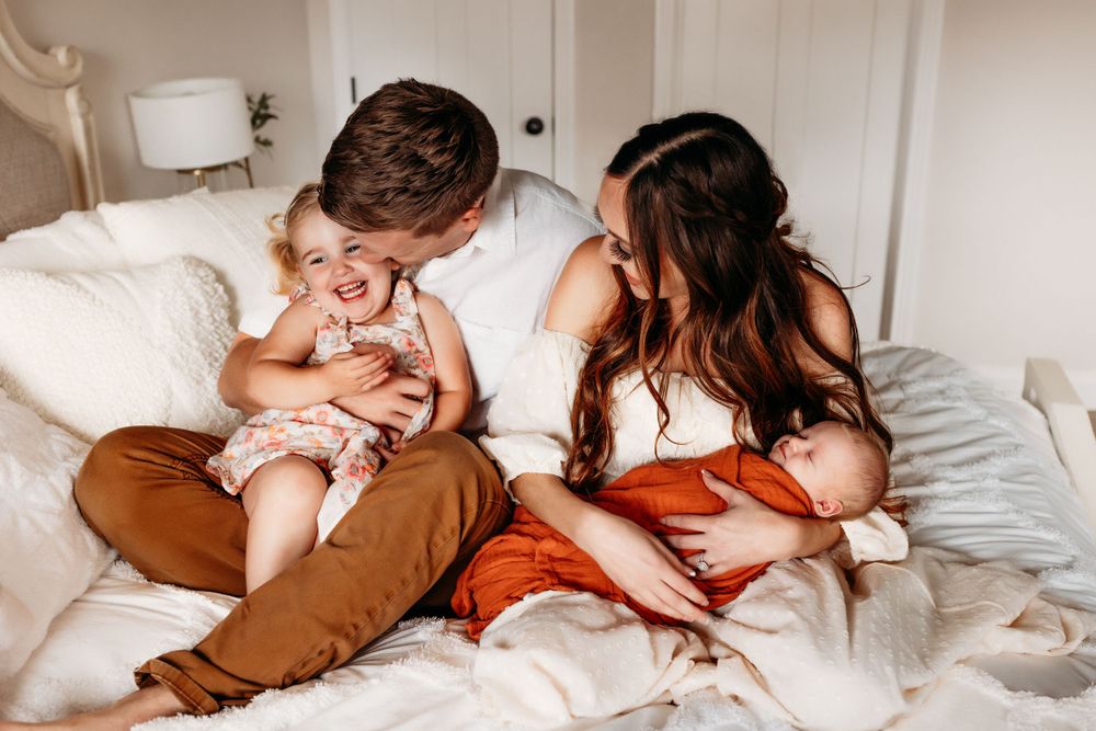 Young couple sitting on white bed laughing with toddler girl and newborn baby.