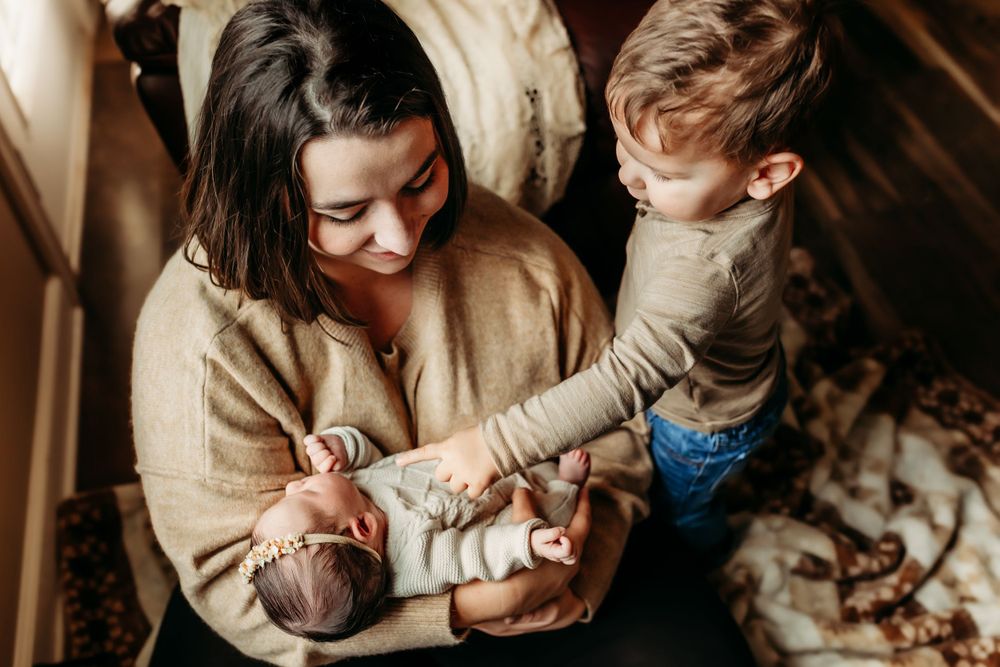 Mother holding newborn baby while curious toddler sibling reaches out to gently touch infant.