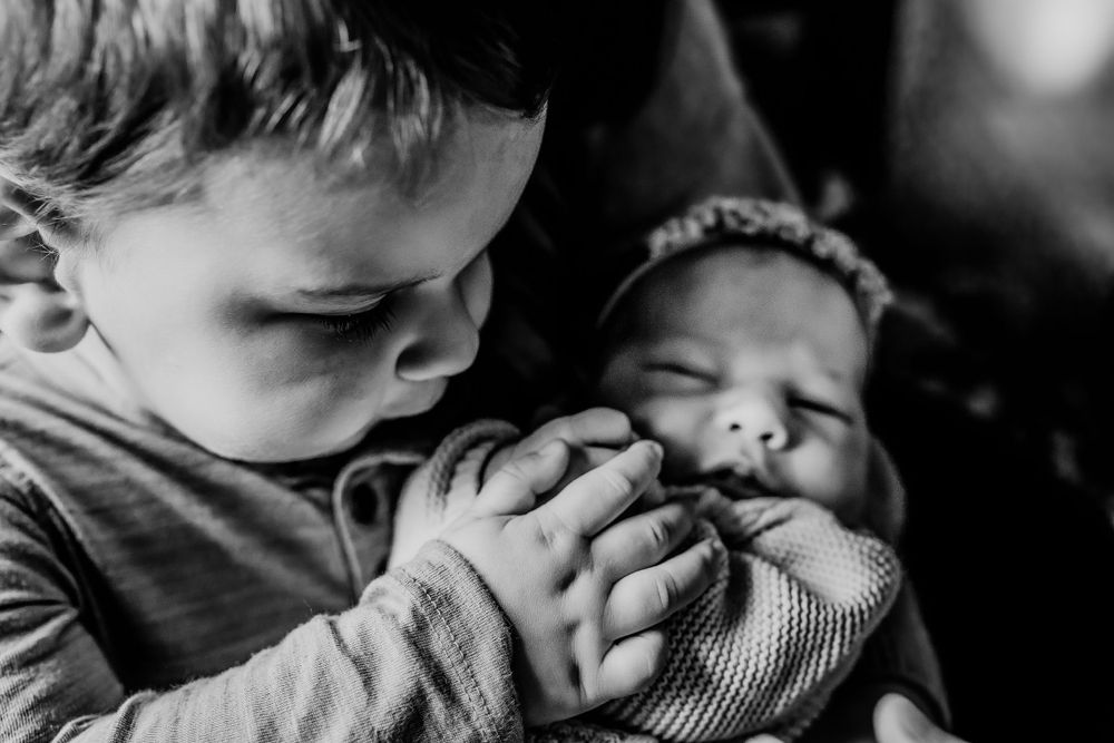 Black and white photo of a young toddler boy gently kissing a sleeping newborn baby sibling.