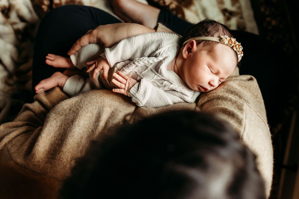 Sleeping newborn baby girl in white outfit with floral headband resting on mothers lap.
