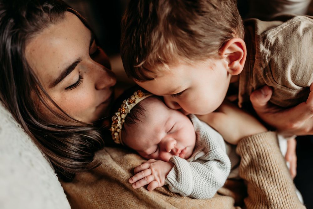 Mother kissing newborn baby while toddler sibling leans in to give a gentle kiss.