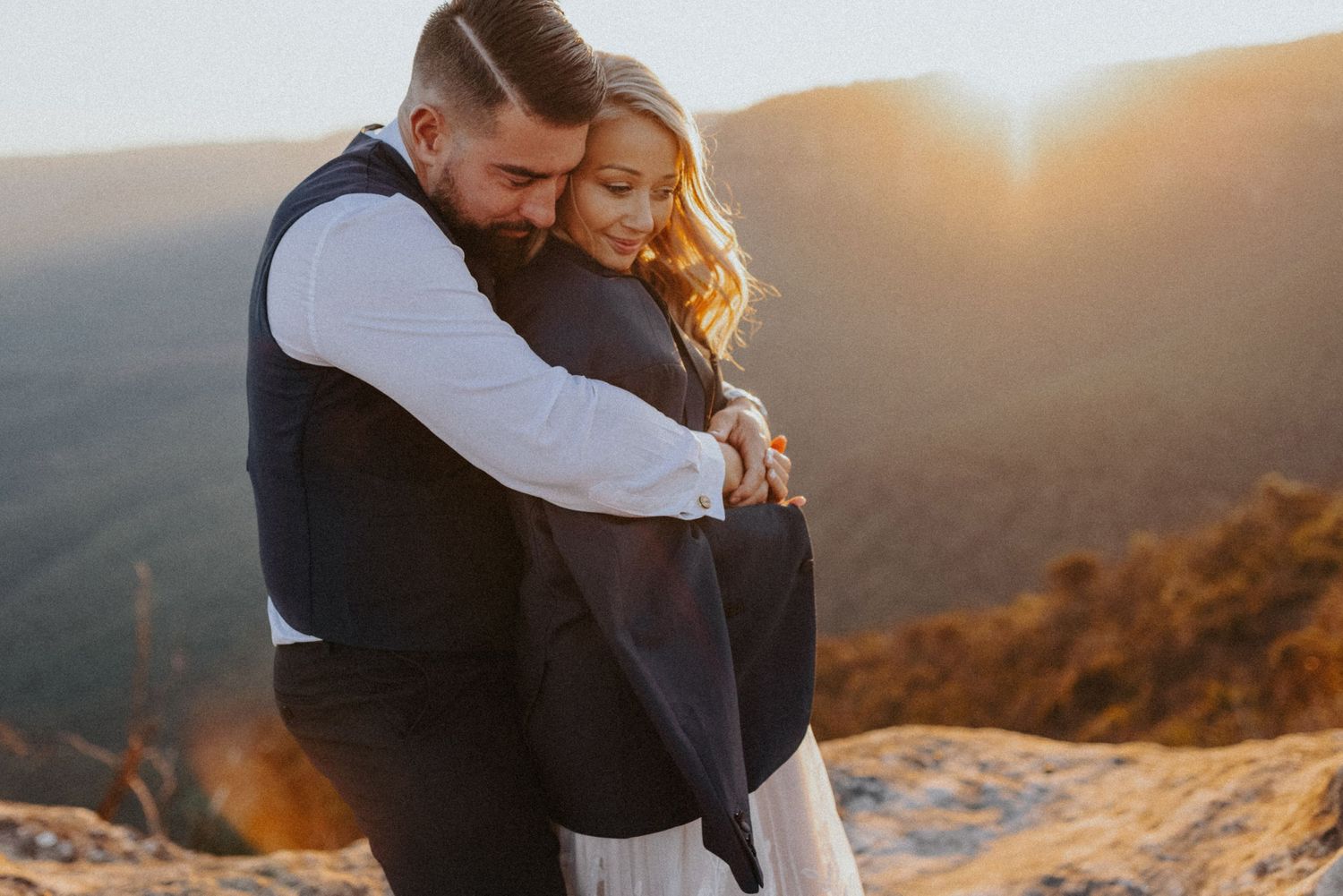 Couple embracing at golden sunset on rocky mountain overlook, bride in white dress and groom in vest.