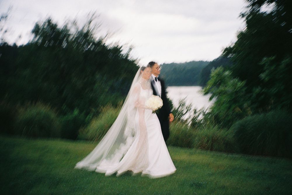 A dreamy wedding photo captured on film shows a couple embracing on a lush green lawn beside a lake at dusk.