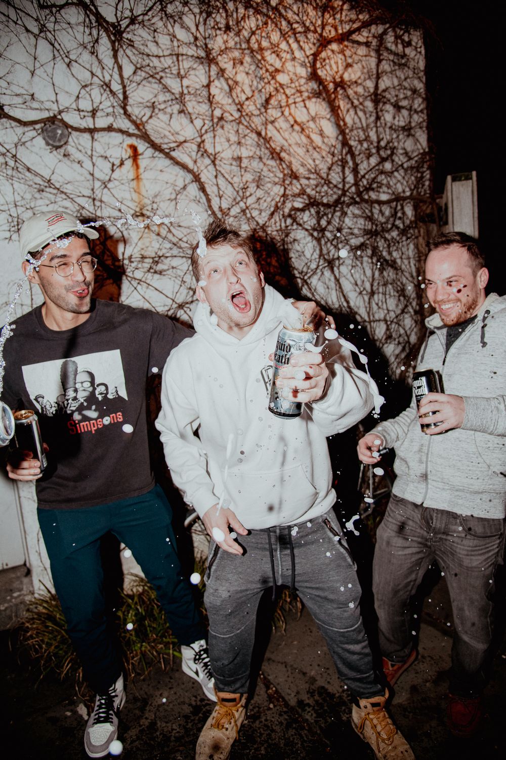 Three young men celebrating outdoors at night with drinks and confetti falling around them.