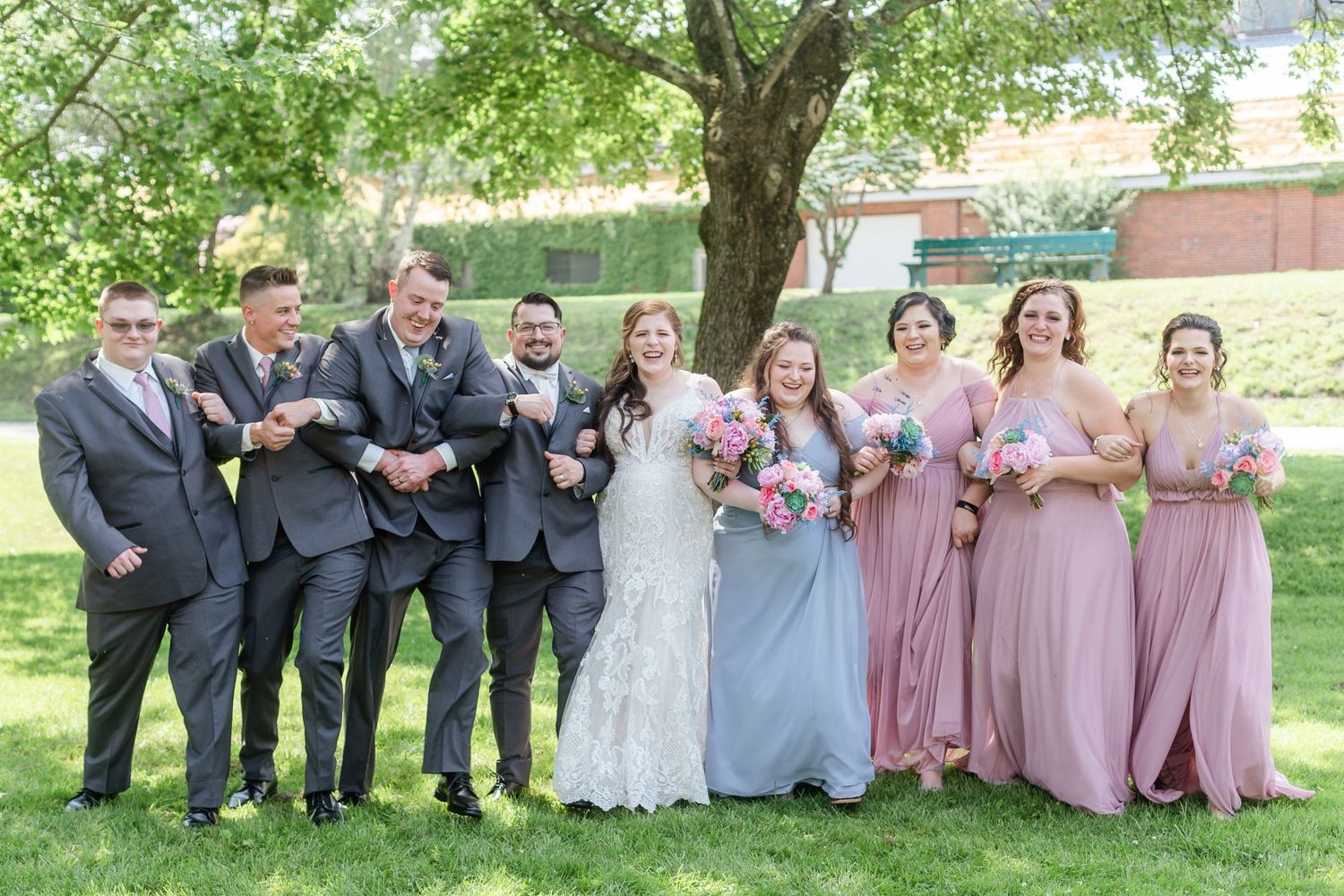 Wedding party in gray suits and mauve dresses poses together outdoors under green trees.