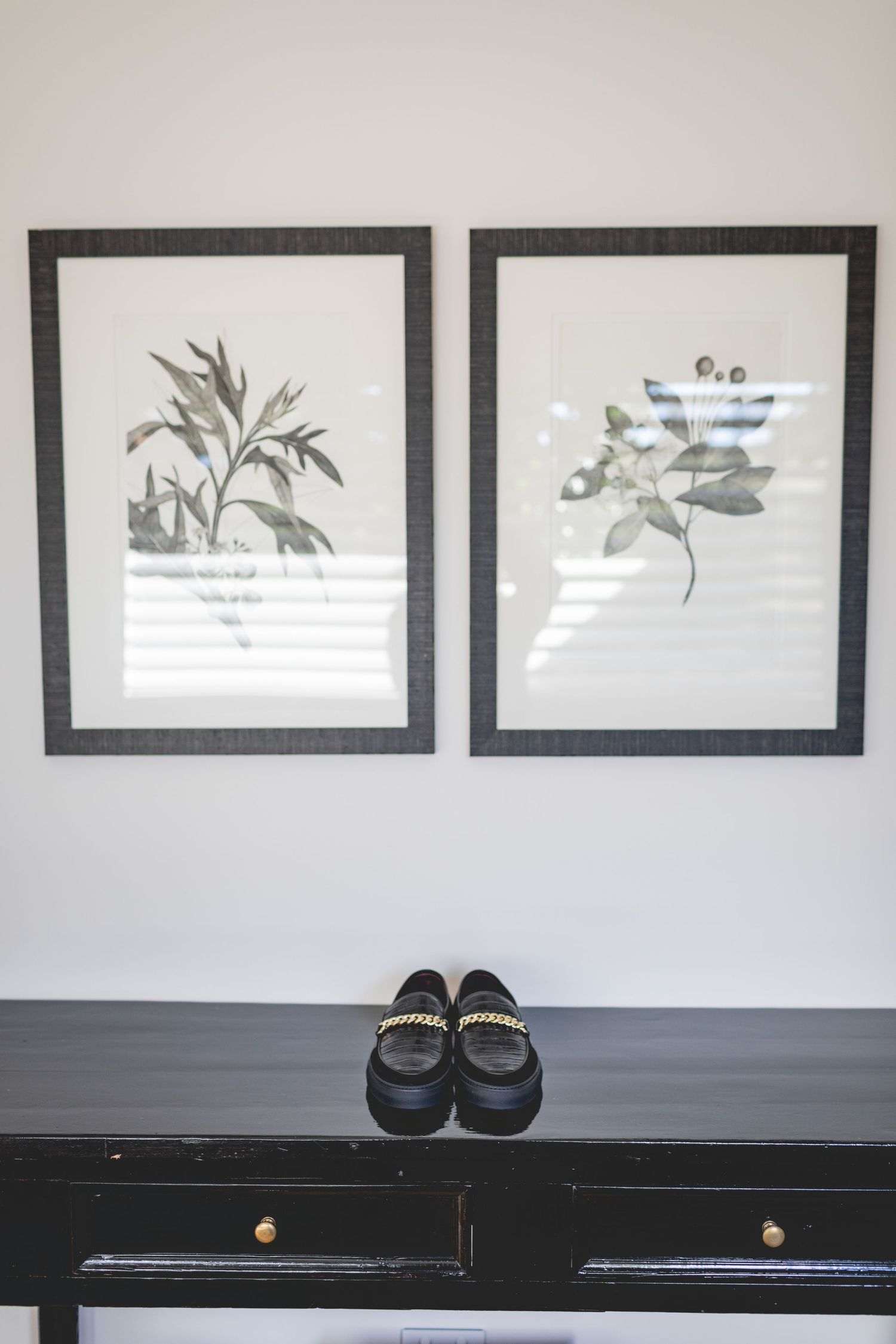 Botanical prints in black frames displayed above loafers on dark wooden console table.