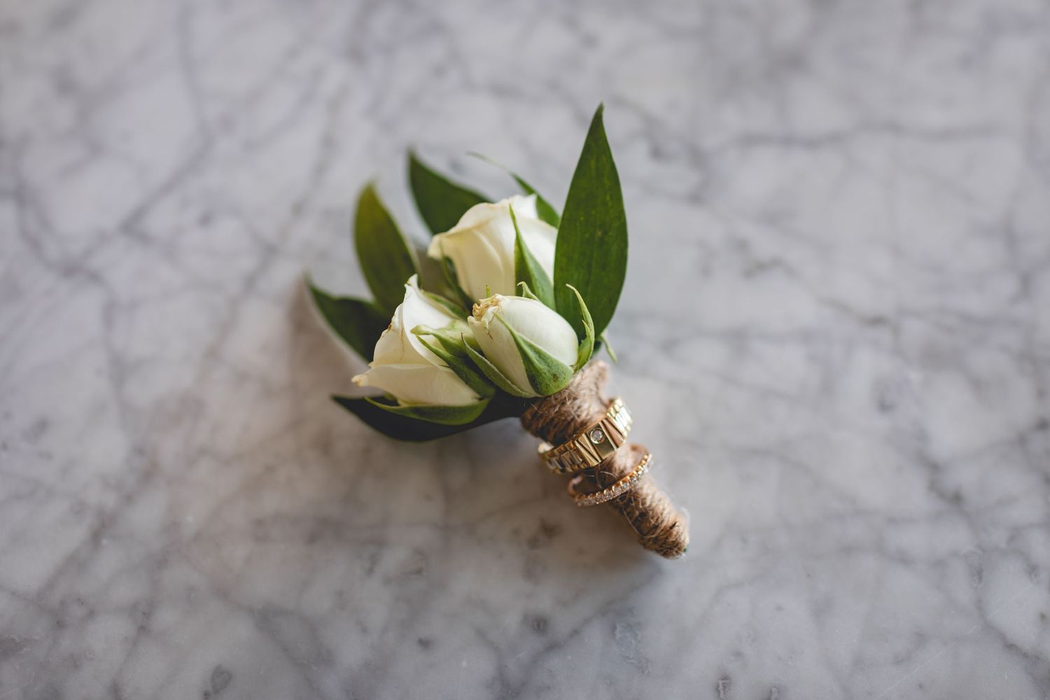 A delicate white rose boutonniere with green leaves wrapped in twine on marble surface.