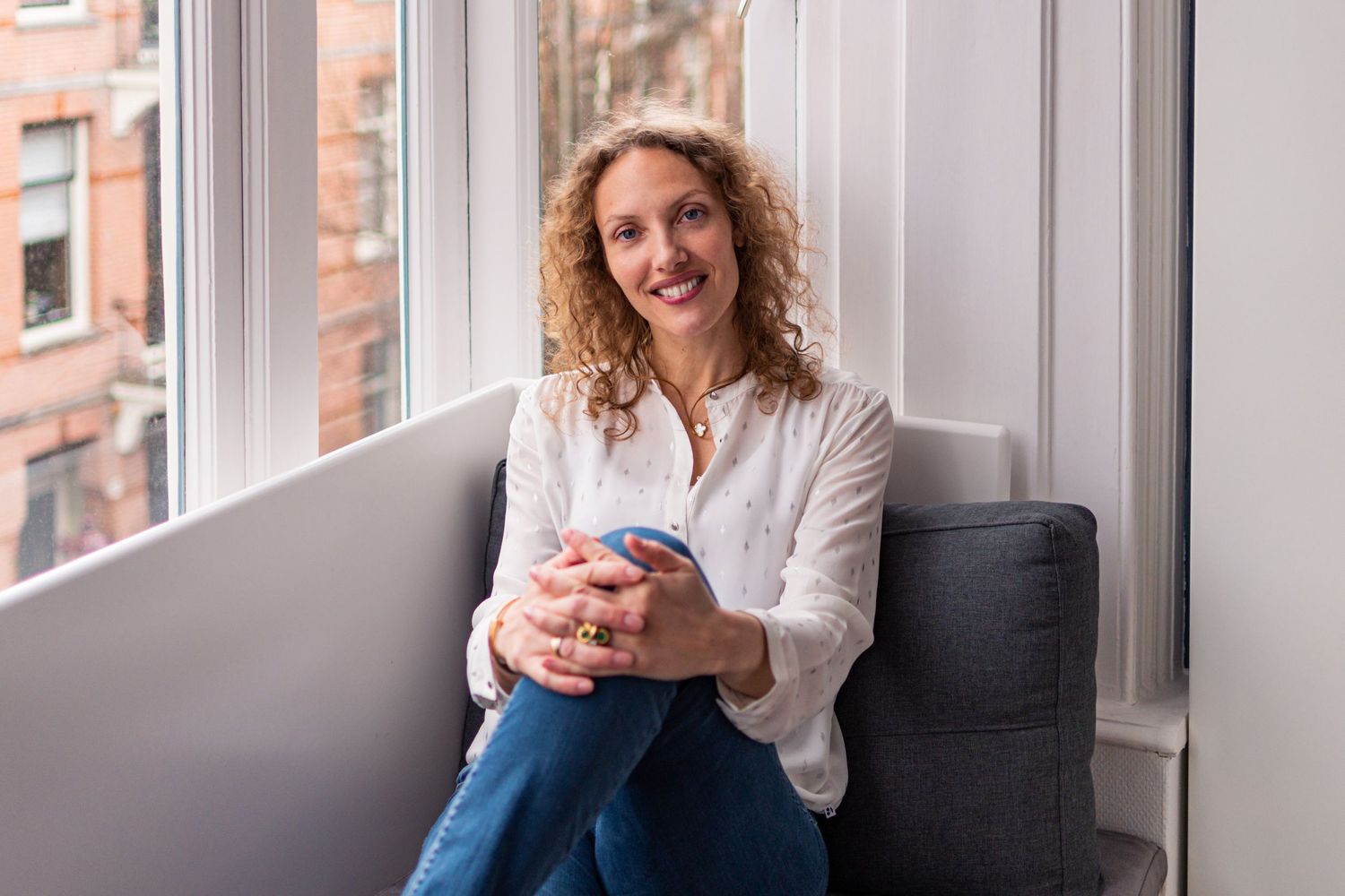 Portrait photo of a smiling woman sitting next to a window at home in Amsterdam and looking at the camera