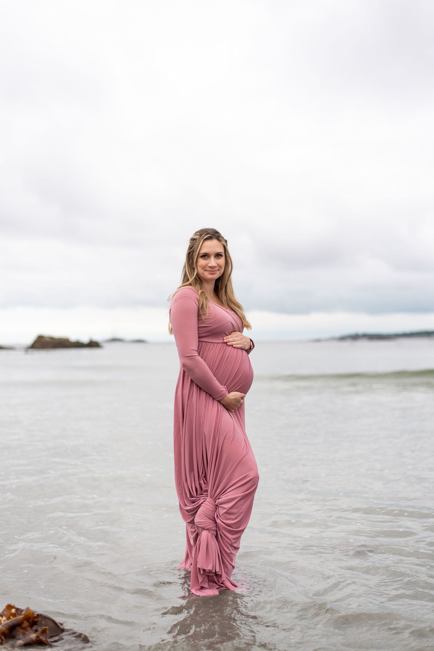 A person in a flowing pink dress stands in shallow ocean water during a cloudy day.