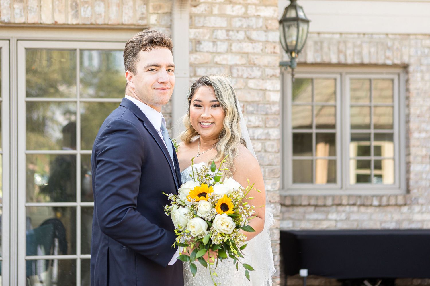 A newlywed couple stands outside a stone building adorned with white flowers and pops of yellow in their bouquet.