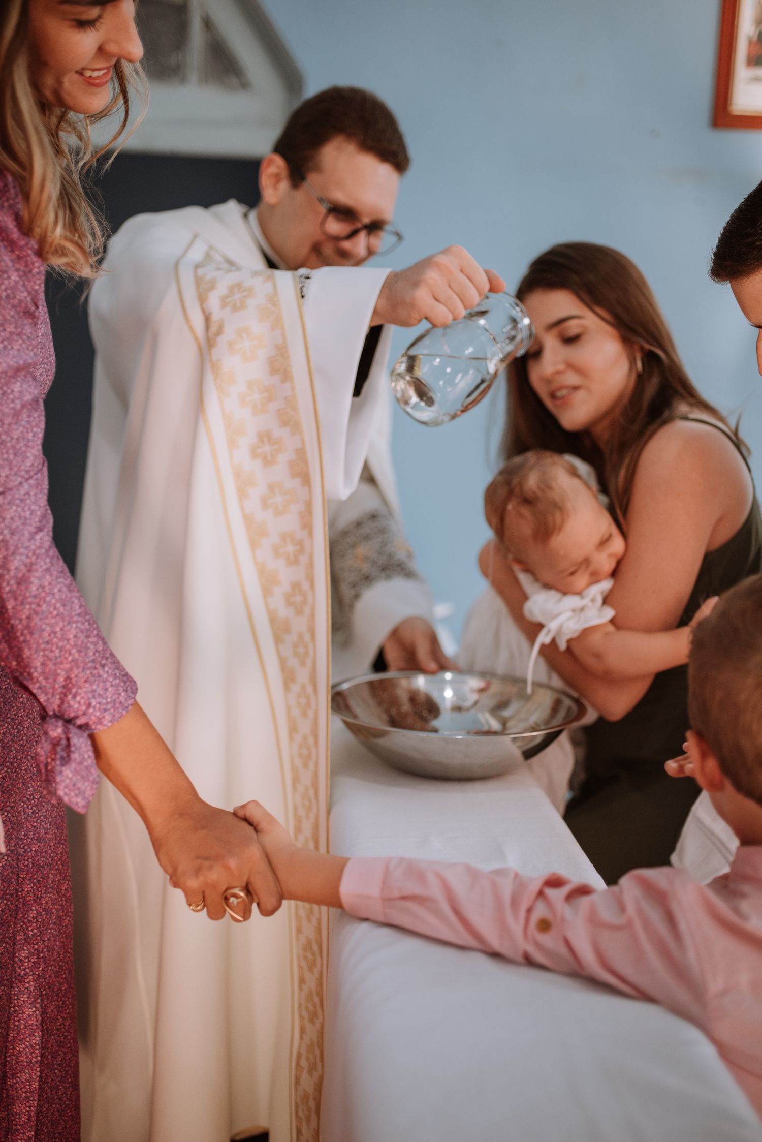Baptism ceremony in progress with priest performing blessing while family gathers around.