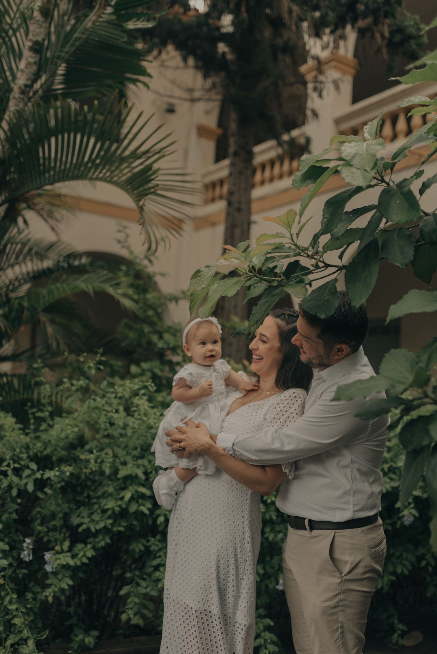 A family in white poses together in a lush garden setting with palm trees and greenery in the background.