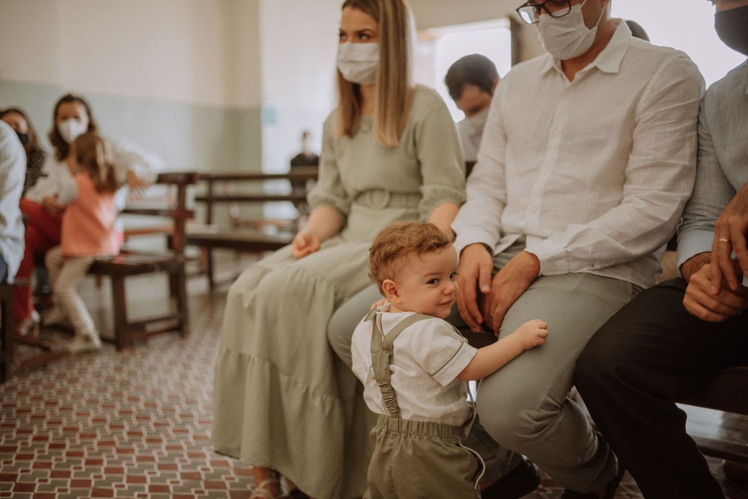 Family gathered on church pew during ceremony with people wearing masks in background.