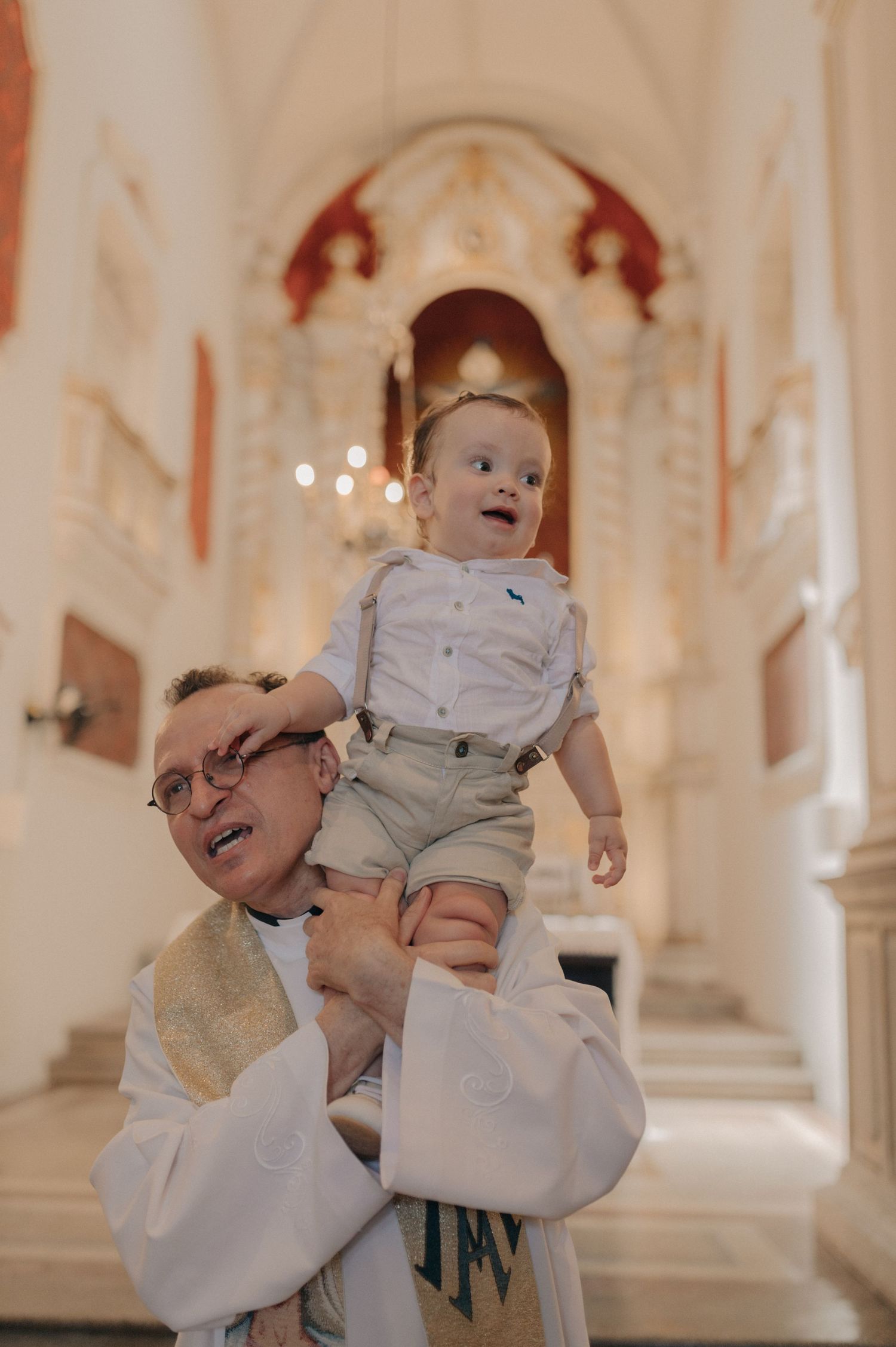 Priest in white robes holds up smiling baby in front of church altar.