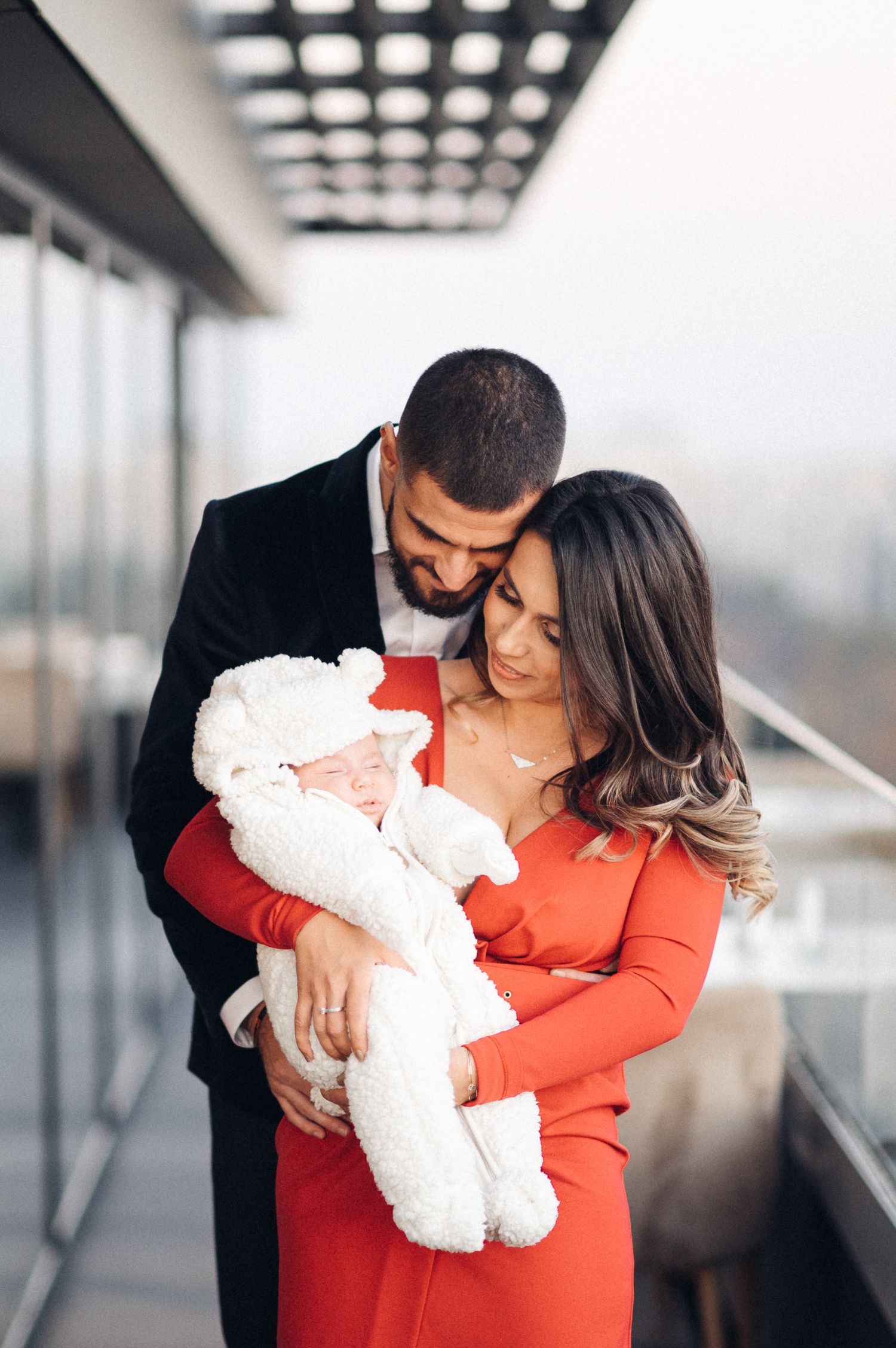 An intimate indoor portrait with a newborn baby wrapped in white, against modern architecture.
