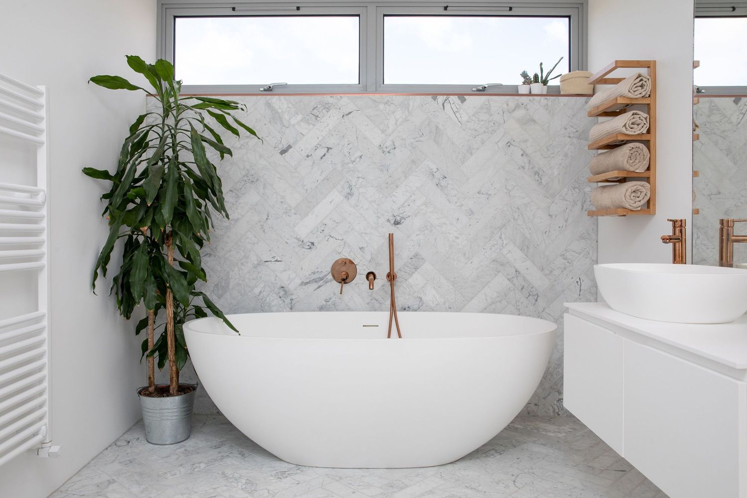 Modern bathroom with freestanding white tub, marble herringbone wall, and tropical plant.