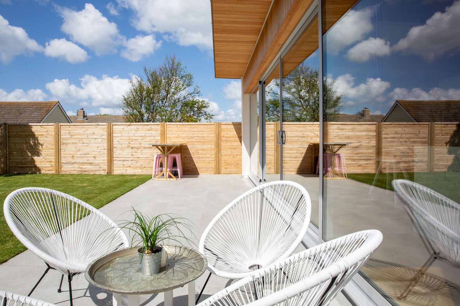 Stylish outdoor seating area with white Acapulco chairs and glass table on a covered patio with wooden fence.