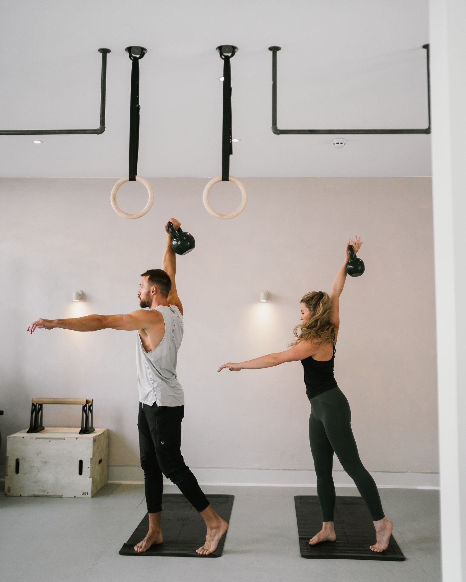 Two people performing synchronized kettlebell exercises in a studio with gymnastic rings overhead.