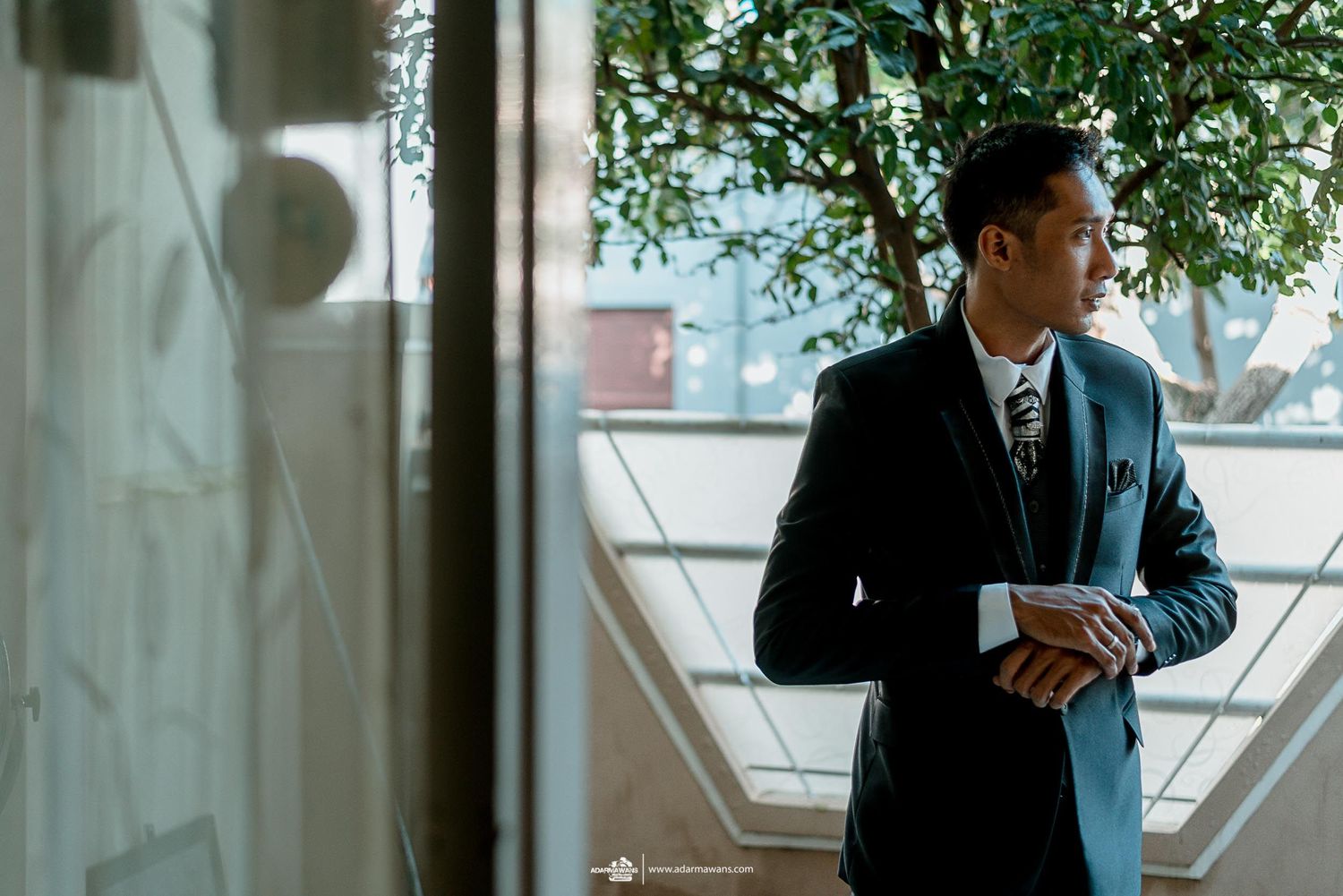 A person in a dark suit adjusts their sleeve near a window with natural light.