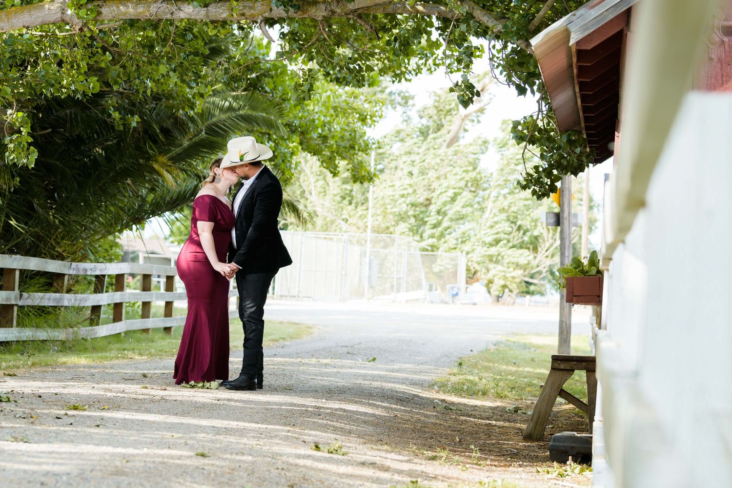 A couple in formal attire embraces on a peaceful tree-lined path with white fencing on a sunny day.