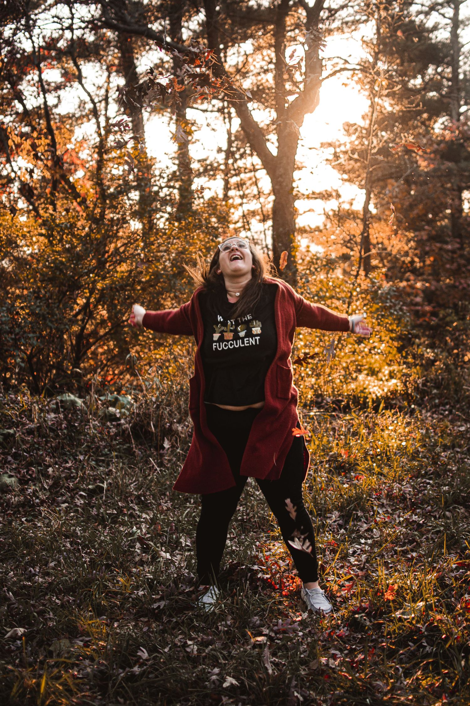 Person in burgundy cardigan poses joyfully with arms outstretched in autumn forest setting.