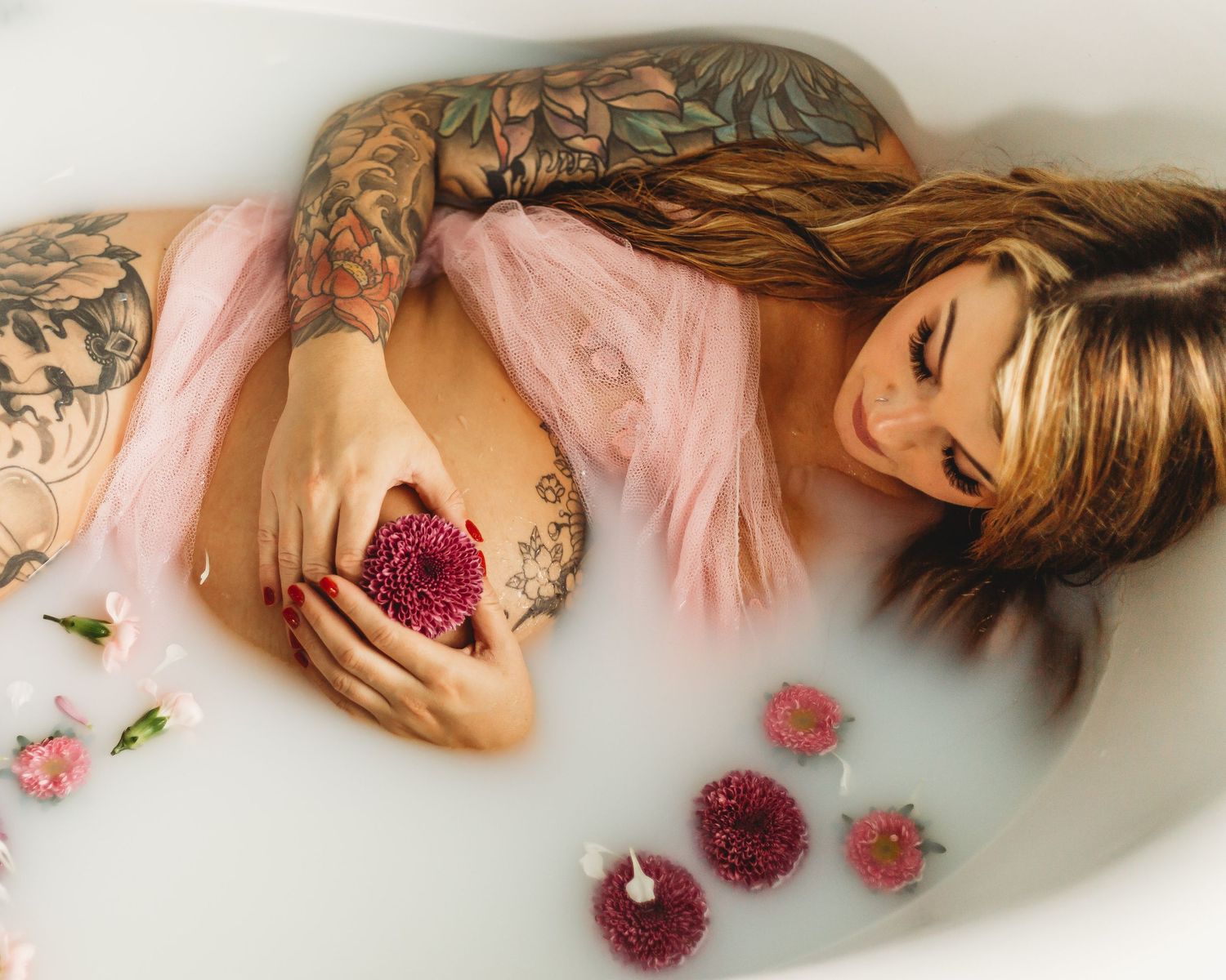 Artistic maternity milk bath photo with pink fabric and burgundy flowers floating in white bathtub.