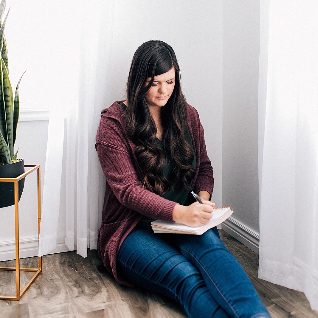 Someone in a burgundy cardigan sits in a corner writing in a notebook with a plant nearby.