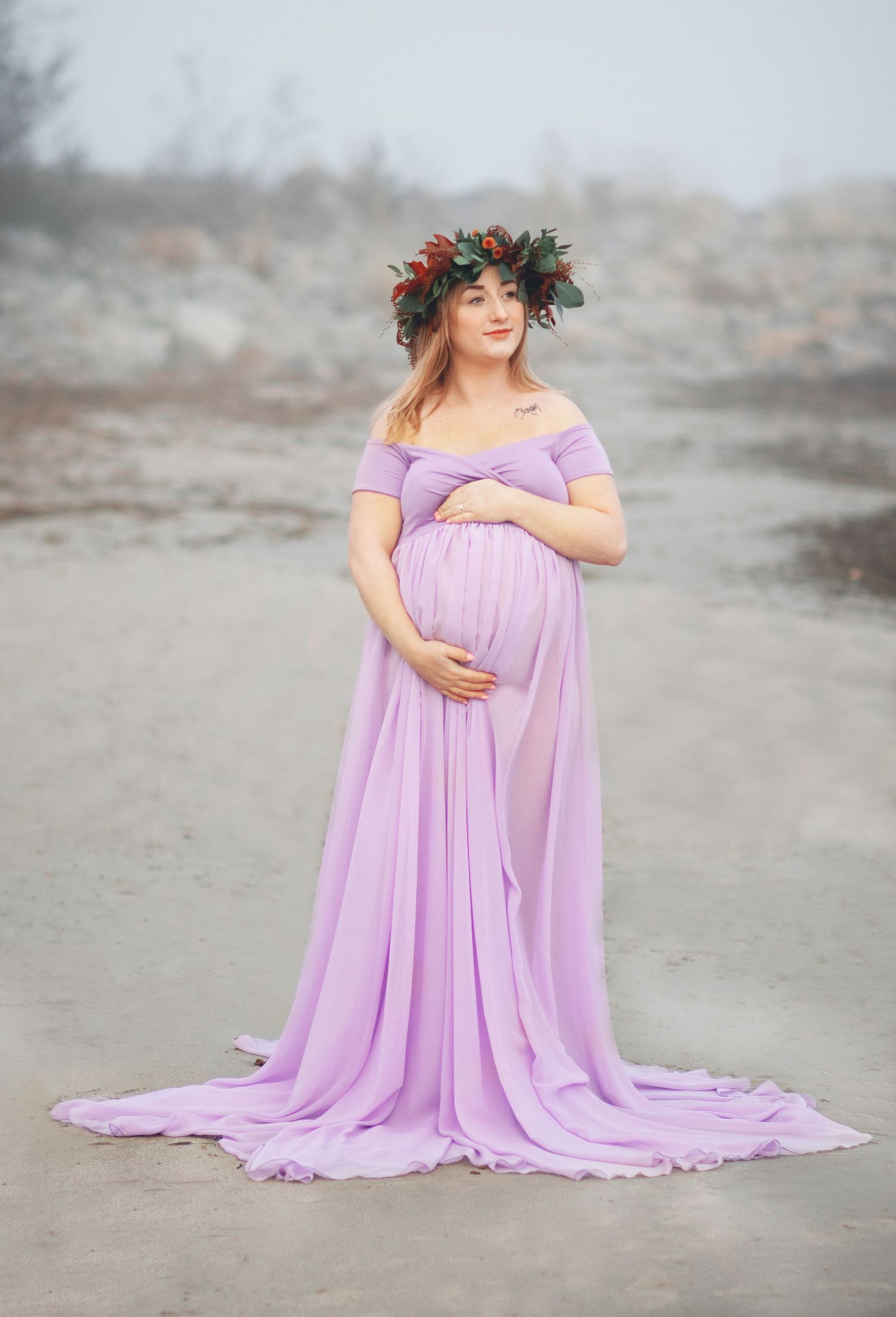 An expectant mother in a flowing lilac dress and floral crown poses on a beach.