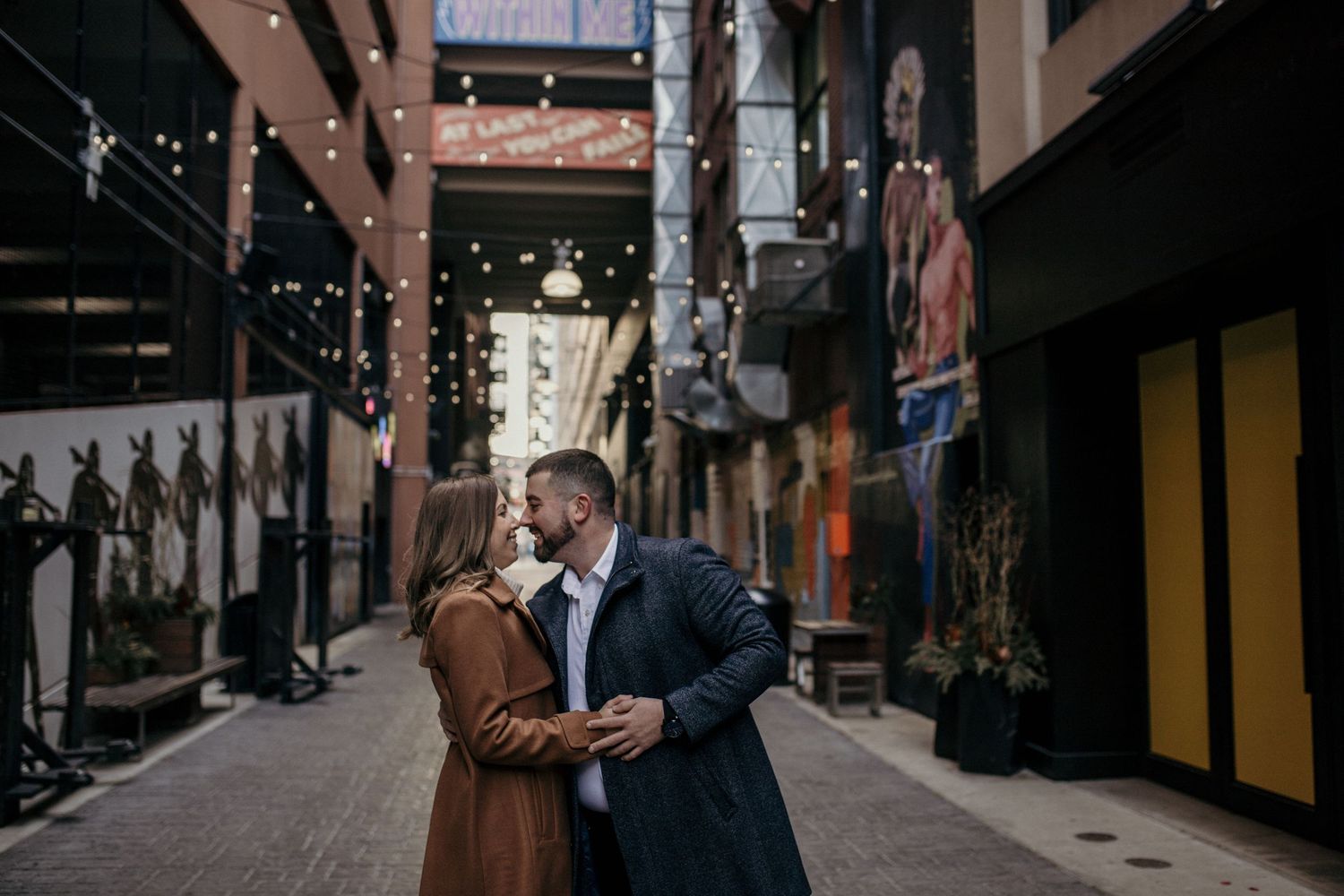 A couple shares an intimate moment in a dimly lit shopping arcade with string lights overhead.