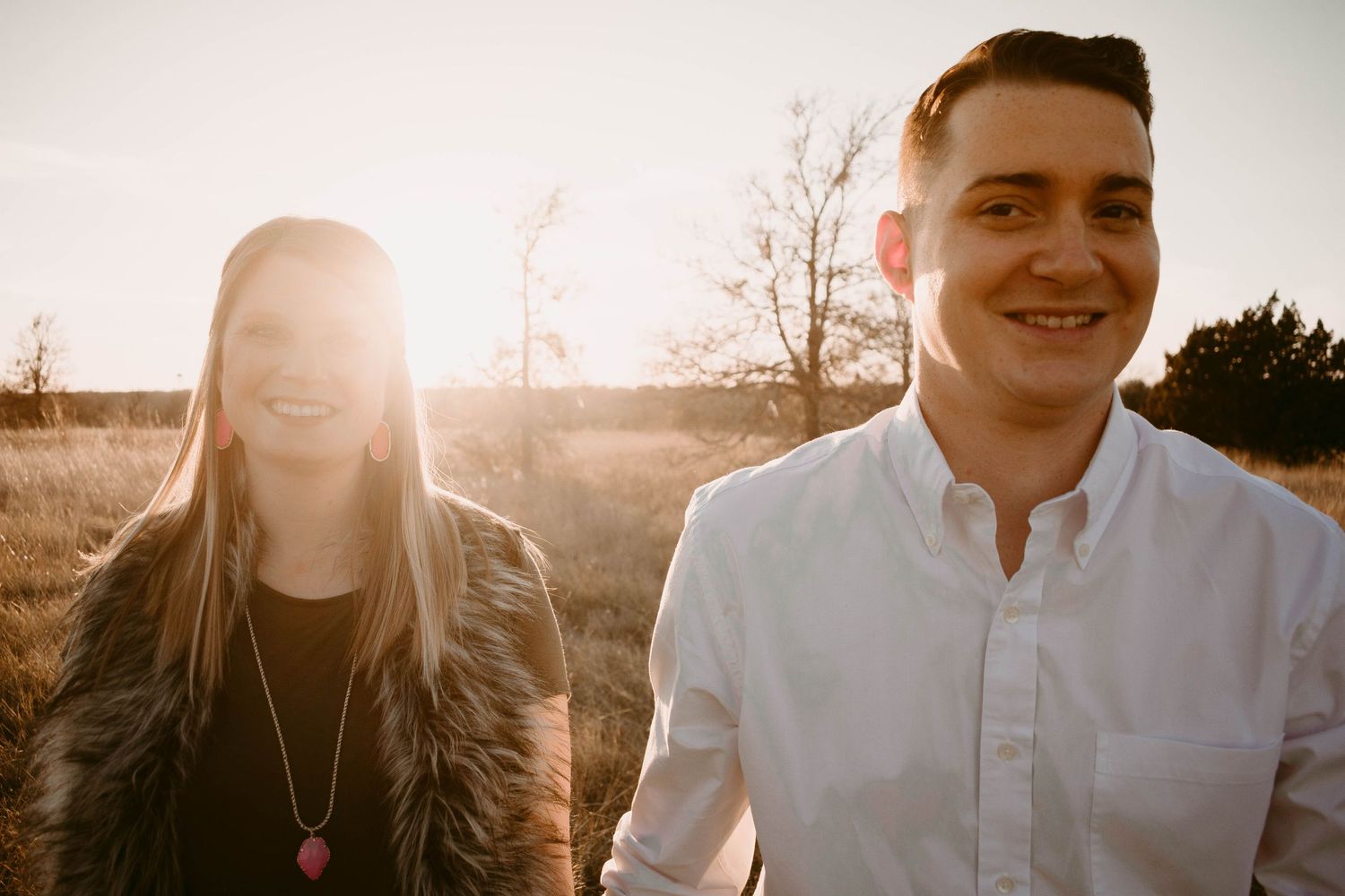 A couple stands together in a field during golden hour with sunlight creating a beautiful backlit effect.