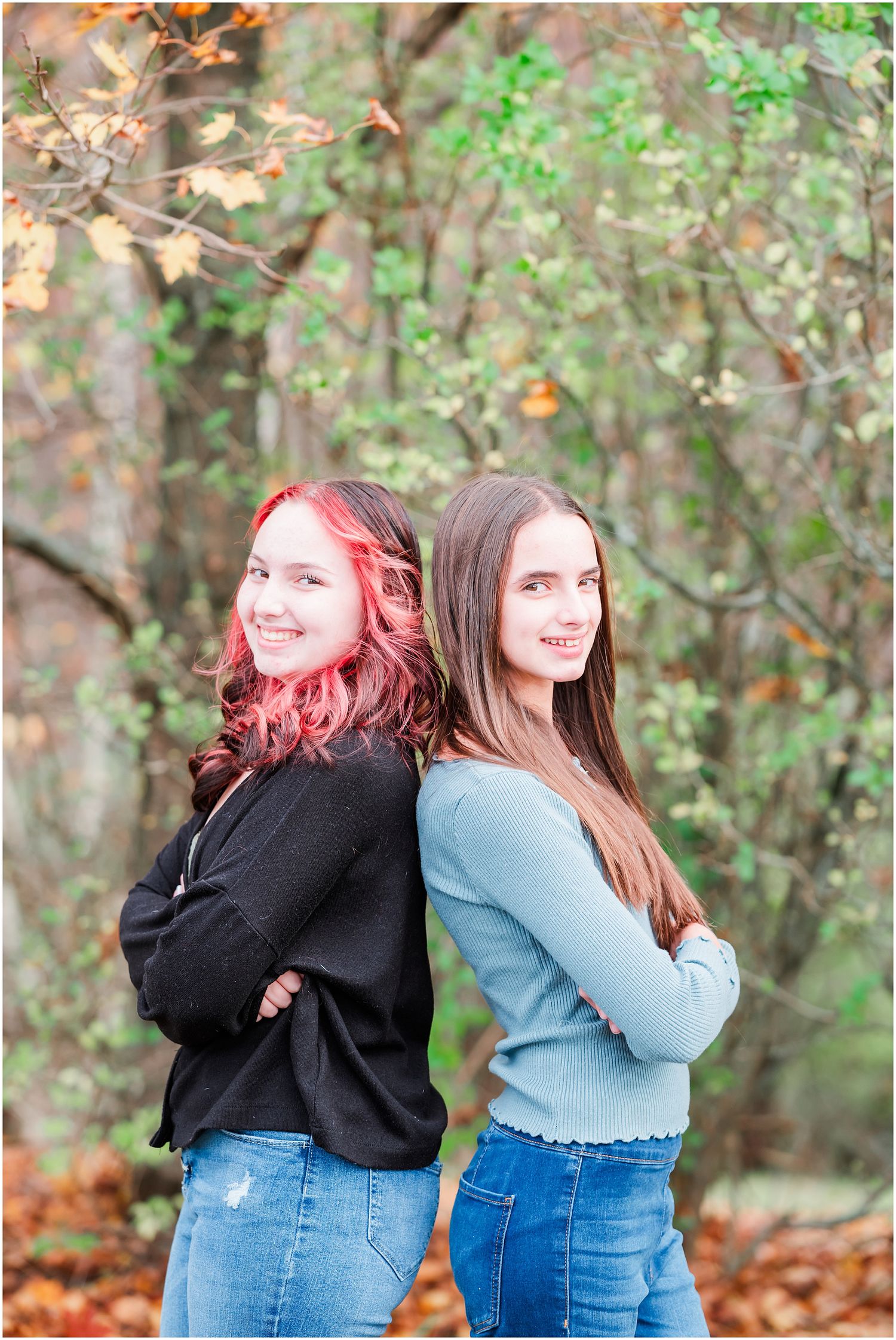Two people pose back-to-back outdoors with autumn foliage in the background.