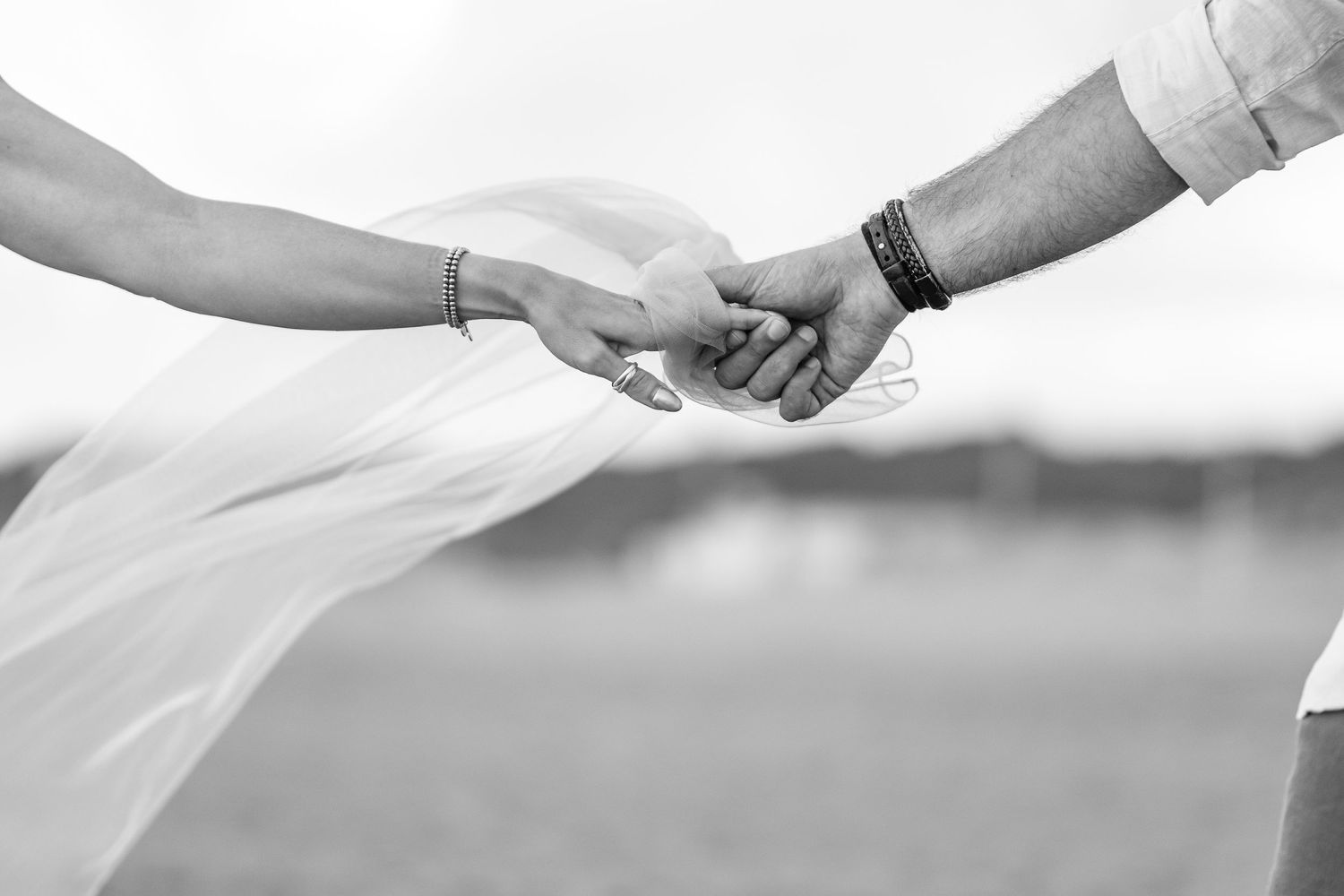 Black and white close-up of two hands clasping together with a flowing wedding veil in the background.