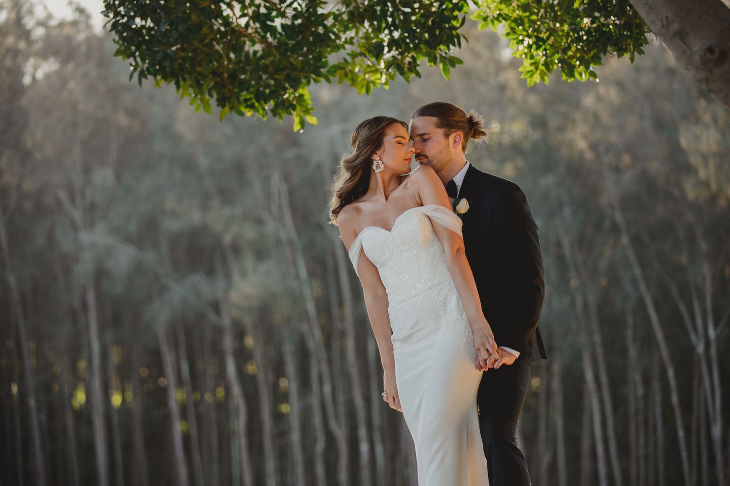 A wedding couple shares intimate moments in a serene forest setting with tall trees.