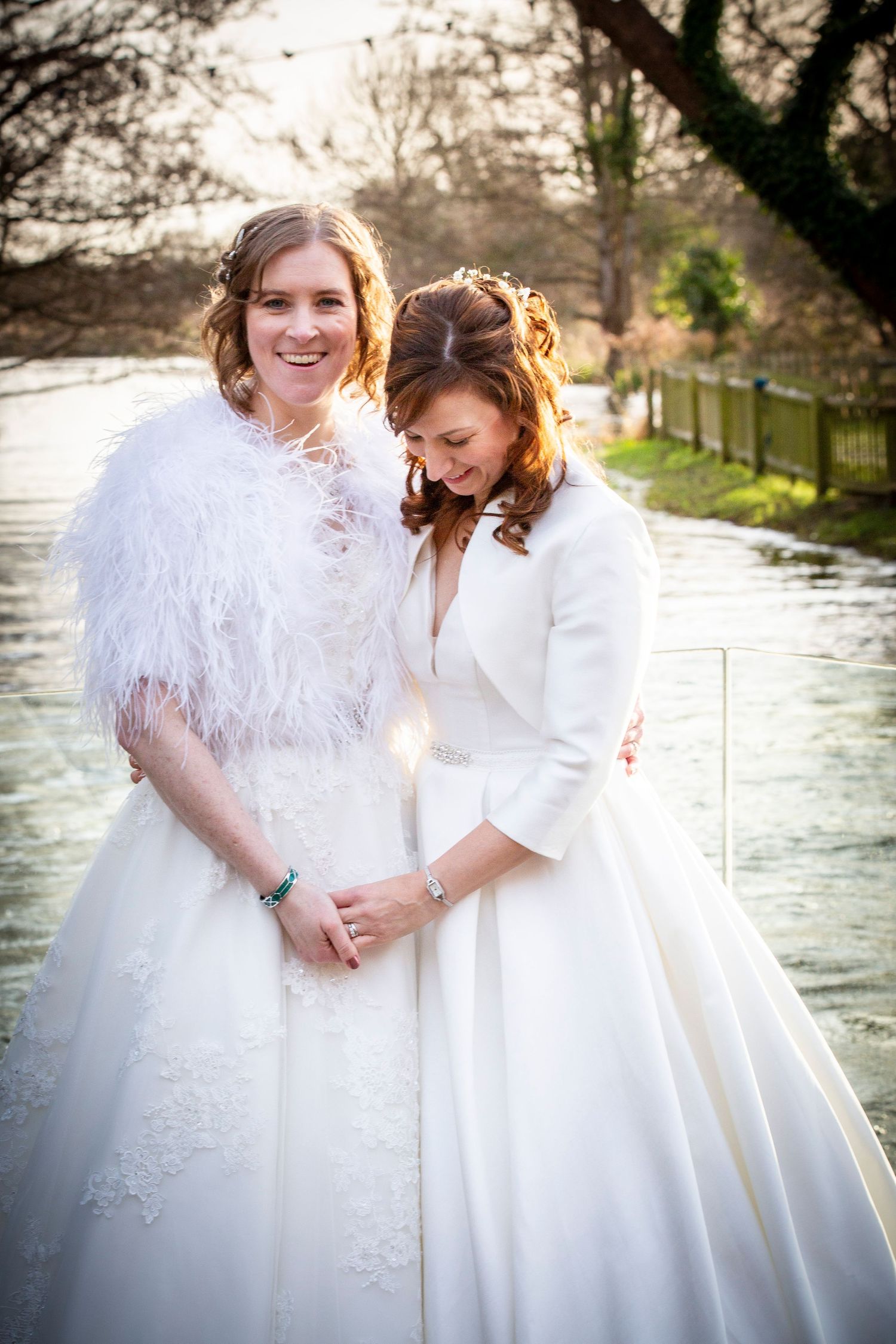 Two brides in white gowns pose by a river, one wearing a dramatic feather wrap.