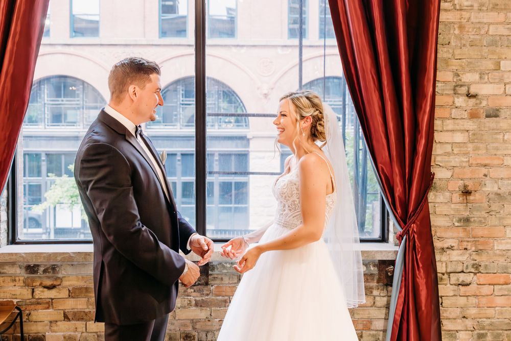 Wedding couple sharing joyful moments by large windows with exposed brick walls.