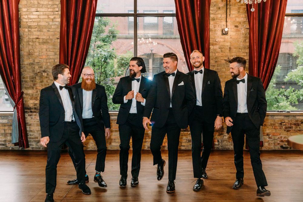 Groomsmen in black tuxedos share a candid moment in an elegant brick-walled venue with red curtains.