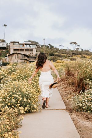 Woman in white dress walking along coastal path lined with yellow wildflowers and houses on hillside.