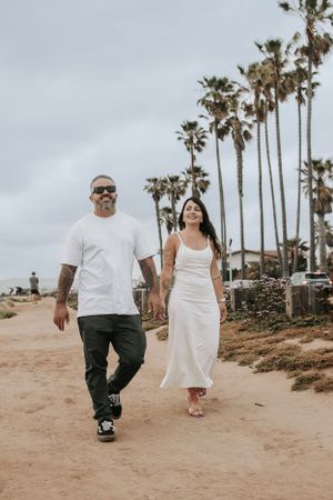 Couple walking together on coastal path with tall palm trees against cloudy sky.