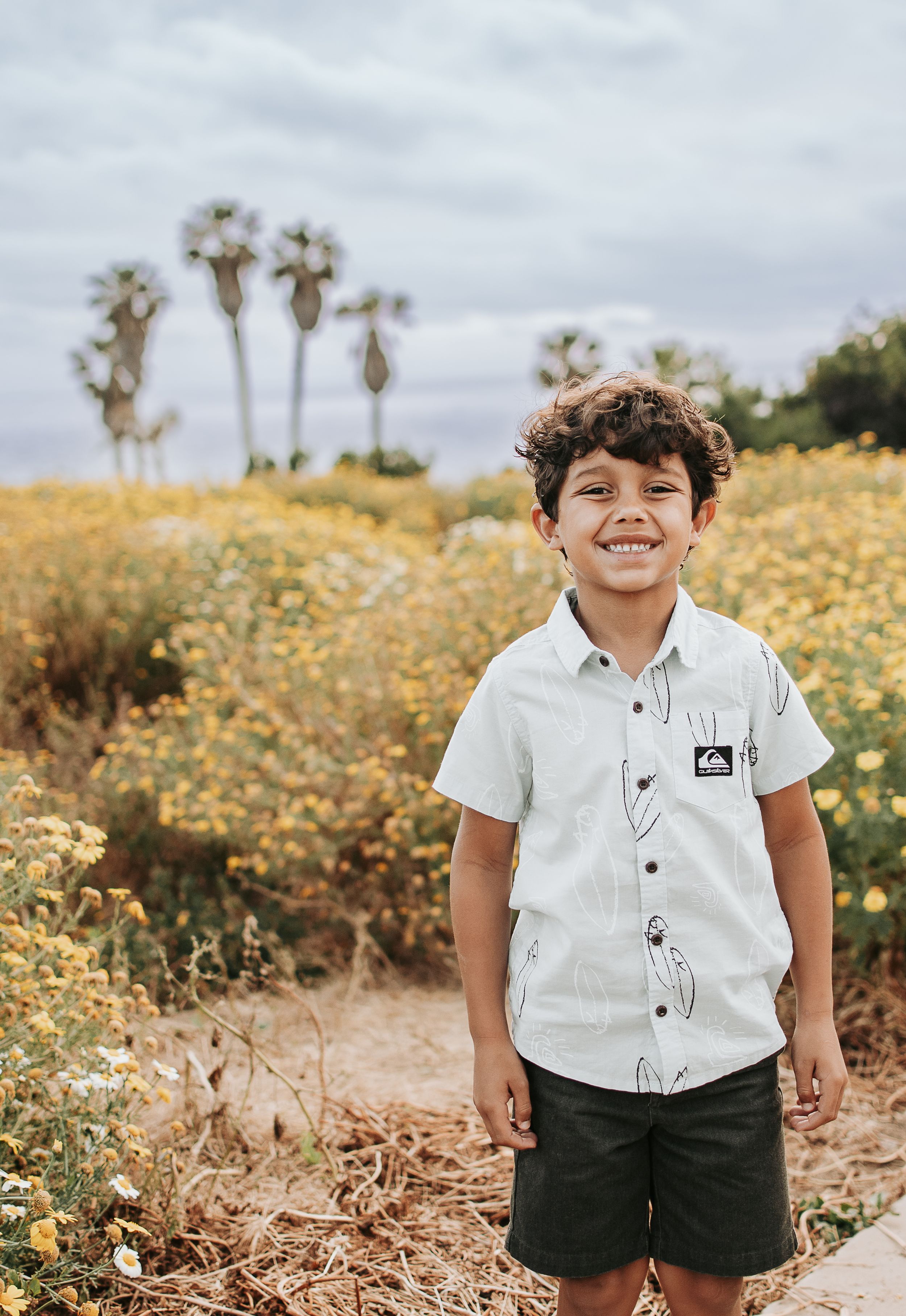 A young boy stands smiling in a white shirt among yellow wildflowers and palm trees.