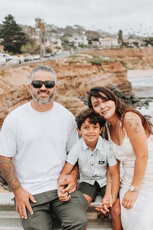 Casual family photoshoot on a sunny day at the beach with rocky cliffs in background.
